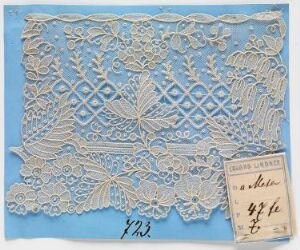  Intricate ivory lacework featuring floral designs and geometric patterns on a blue background, accompanied by a tag with handwritten text, possibly detailing the lace's origin or maker.