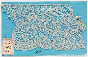  An intricate piece of white lace with floral designs, pinned to a bright turquoise background, with a small paper tag bearing handwritten text attached to the lace.