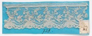  An elongated piece of intricate white lace with a floral pattern displayed against a cyan background, accompanied by a small tag with handwriting in the lower right corner.