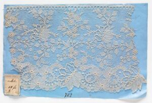  A piece of antique floral patterned lace on a light blue background with a small, aged paper tag attached to the bottom left corner, displaying faint handwriting.