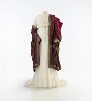  A historical gown reminiscent of ancient Roman or Greek attire on a mannequin, featuring a creamy white fabric with a deep red or burgundy draping cloak, displayed against a simple, light-colored background. Artist name and title are unknown.