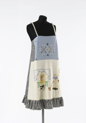  A mannequin displaying a multi-fabric dress by Julie Skarland, featuring a blue upper section with hand-embroidered beads, a central colorful embroidered rectangle, and a lower section with a black and white checkered ruffle.