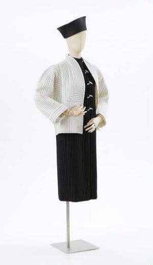  A mannequin dressed in a classic design by Per Spook, featuring a white textured blouse with puffed sleeves, a black vest or sash with white detailing, and a knee-length black skirt, accompanied by a small, flat-topped black hat. The outfit blends monochromatic sophistication with a touch of vintage elegance.