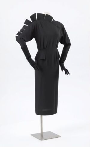  A black knee-length dress with tiered shoulder detailing and fitted waist, displayed on a female mannequin. The dress, designed by artist Per Spook, is complemented by black leather gloves, emphasizing a sleek and modern style.