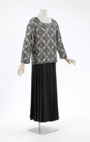  A mannequin displays a two-piece outfit by Per Spook, consisting of a black and white geometrically patterned blouse with long sleeves and a scoop neck, paired with a long, flowing black skirt. The ensemble epitomizes elegance with its sharp contrasts and flowing lines.