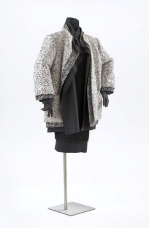  A fashion ensemble designed by Per Spook, featuring a textured white and gray jacket over a flowing black undergarment, displayed on a mannequin against a neutral background. The outfit illustrates a sophisticated interplay of color, texture, and form, with both machined and hand-sewn elements in silk fabric.