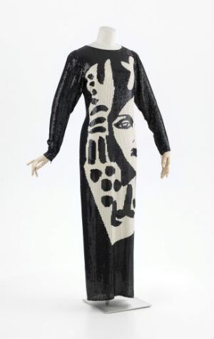  A mannequin displaying a black and white silk chiffon full-length gown titled "Place de l'Étoile: Noir - Blanc, nr. 20" by Per Spook. The dress features abstract white patterns resembling facial features embellished with shimmering plastic sequins against a contrasting black backdrop, with sheer black sleeves and elegant floor-length design.