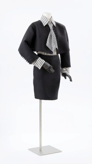  Sophisticated black and white outfit designed by Per Spook, featuring a short-sleeved puff-shoulder blouse, a knee-length skirt, a striped scarf, a checkered band hat, and black leather gloves against a white background.