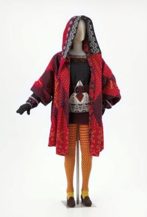  A mannequin without facial features dressed in a colorful hooded outer garment with intricate patterns in deep purple, black, red, and white, layered over a black patterned shirt and vest, with an orange-red striped skirt, yellow-orange tights, and black and yellow shoes against a plain light background.