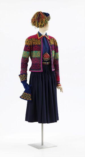  A mannequin is displayed wearing a colorful, patterned ensemble by Per Spook, featuring a multicolored knitted hat, a patterned cropped jacket in green, red, purple, and yellow, a dark blue silky scarf, a navy pleated skirt, and matching gloves, all incorporating various materials and techniques.