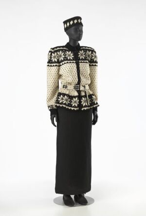  Mannequin dressed in an Ellinor Flor knit costume with a cream and black patterned jacket, featuring geometric and floral motifs, black skirt, and matching cap against a light background.