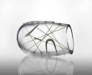  "Mønster" by Ina Kristine Hove, an abstract blown glass art piece with metallic wire details creating a geometric pattern within its semi-opaque, irregular shape, set against a light background with a reflective surface below.