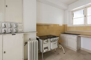  Historical kitchen design "Im Burgfeld 18 (Frankfurter Küche)" by Margarete Schütte-Lihotzky, featuring an off-white color scheme with a gas stove, tall cabinet, radiator, wood countertops, and a well-lit double window.