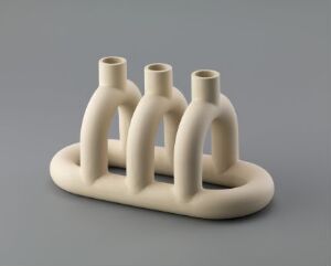  The "Kapp lysestake" by Noidoi (Kathrine Lønstad and Cosmin Cioroiu), a minimalist beige ceramic candle holder with three interconnected tubular holders on a single base, displayed against a soft gray background.