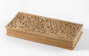  Intricately carved wooden box with floral patterns on a plain off-white background. The box is rectangular with delicate carvings on all sides, displaying a warm light brown tone and textured ornamentation.