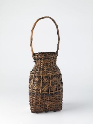  A handwoven brown basket with a darker brown handle and geometric patterns, set against a light gray background. The artist's name and title are unknown.