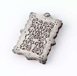  An intricate silver filigree pendant with scalloped edges and a complex lace-like pattern, showcasing floral motifs against a concealed deep red underlayer, featuring a loop at the top for hanging.