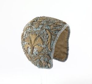  An ornate historical cap with intricate embroidery in shades of gold, olive green, muted blues, and browns on a creamy fabric, lined with dark brown fur or velvet, showcased against a plain white background.