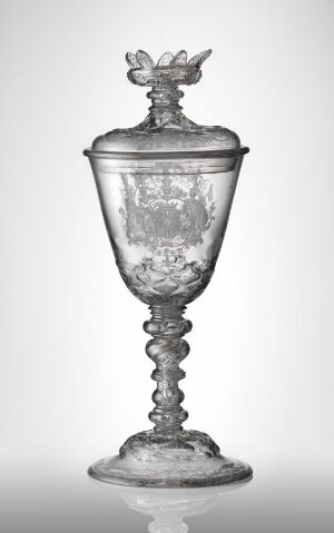  An elegant glass goblet with a lid, featuring an ornate bird figure atop, intricately etched designs on the bowl, and a twisted spiral stem, on a graduated light to dark background.