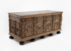  An intricately carved dark brown wooden chest with high-relief panels and black bulbous feet stands against a neutral background, highlighting the detailed craftsmanship and historical design of the piece.