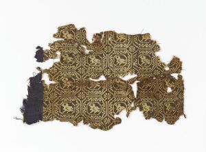  An ancient, ornate golden-brown textile with intricate geometric and floral patterns, displayed against a light background, showing signs of wear with multiple holes and frayed edges. The artist and title are unknown.