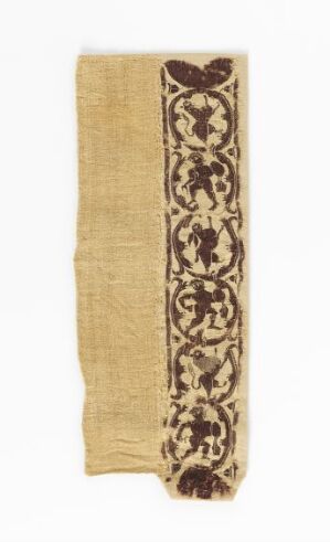  A long, narrow, beige textile fragment with a dark brown intricate symmetrical pattern running down the center, featuring vine-like designs and alternating floral emblems, showing signs of age and wear. Artistname and title are unknown.