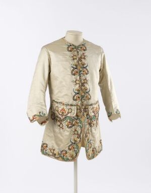  A mannequin presenting an 18th-century style cream-colored jacket and breeches set, ornately decorated with a symmetrical floral motif in earthy green, brown, and muted red tones.