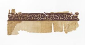  A fragment of aged material with a textured beige to light brown background and a dark brown decorative border featuring a repeating pattern that may be floral or vegetal in nature. The fragment has frayed edges and some tears, indicating it is a remnant of a larger antique piece.