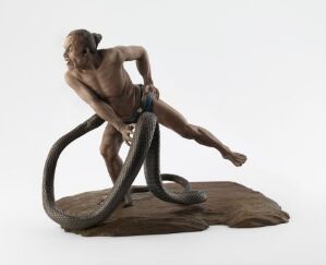  Sculpture of a muscular human figure in a dynamic struggle with a large snake, both cast in varying shades of brown and mounted on a rough, natural-looking base. The man appears to be seizing the snake by its body, his facial expression intense with exertion.