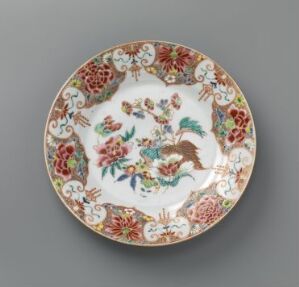  An ornately decorated ceramic plate with scalloped edges, featuring a colorful floral border and a central illustration of two birds amidst flowers. The plate is set against a neutral gray background. Artist name and title are unknown.