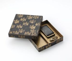  An ornate, dark rectangular box with a gold floral pattern, partially open to reveal a golden-tan interior and a small metallic inkwell, set against a white background.