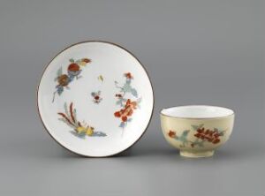  A porcelain cup and saucer set with delicate floral patterns in orange, blue, yellow, and green, displayed against a grey background. The saucer lies flat while the cup is shown at a tilt, revealing part of the interior.