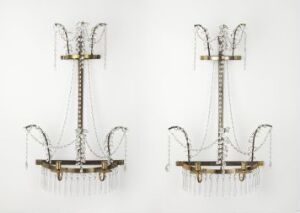  A pair of symmetrical wall fixtures with golden or brass-colored metal details and elongated hanging crystal or glass pendants against a light background, exuding opulence and intricate design.