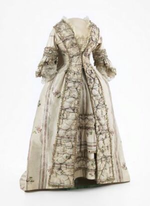  A historical off-white full-length gown with a fitted bodice, tapered puffy sleeves, and a wide skirt, all featuring a delicate pattern of gray or faded black interconnecting branches and floral motifs.