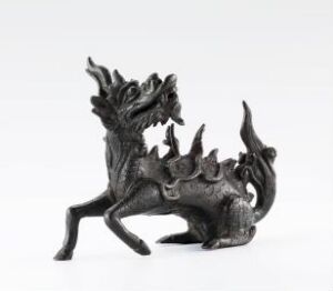  A small, dark gray metallic sculpture of a mythical dragon with an open mouth, detailed scales, and a spiky backbone, set against a light gray background.