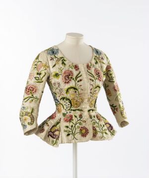  A historical garment with a floral design, featuring long sleeves and a flared waist, displayed on a dress mannequin against a plain background. Colors include a pale cream fabric with red, pink, yellow, blue, and purple flowers, accented with shades of green leaves and hints of gold.