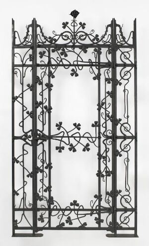  A black and white photograph of an intricate wrought iron gate with symmetrical floral or foliate designs, featuring a complex crest at the top and a stark contrast with a plain white background. Artistname and title are unknown.