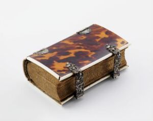  An antique-style object designed to look like a thick book, with a tortoiseshell patterned cover and gilded edges, accented by ornamental silver metalwork at the corners, displayed against a light gray background.