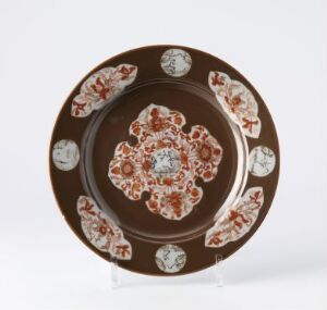  A decorative ceramic plate with a dark brown rim featuring six circular medallions, a lighter brown background, and a central red, green, blue, and white floral mandala pattern.