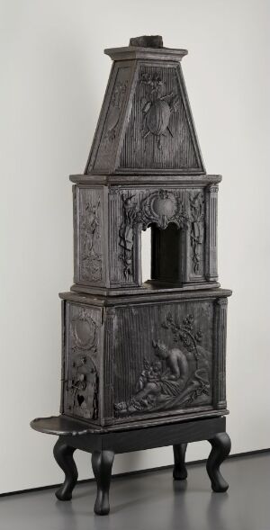  "Støpejernsovn" - an antique cast iron stove with ornate relief designs from Bærums Jernverk, featuring a series of vertically stacked sections sitting on four legs, showcasing the historic merger of utility and artistic craftsmanship in heating appliances.