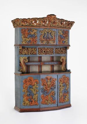  A tall, ornate cabinet with muted blue-grey coloration and panels decorated with vibrant orange, light blue, dark blue, reddish-brown, and gold in swirling patterns and floral motifs. The top section flares out with intricate cut-outs and the entire piece appears to evoke a sense of historic craftsmanship.