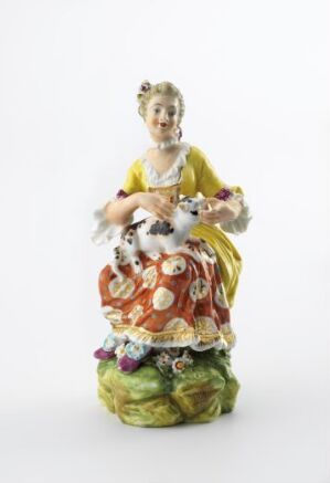  A porcelain figurine of an elegant 18th-century woman with a yellow and red floral dress, holding a small white dog and surrounded by flowers and additional dogs on a green grass-like base.