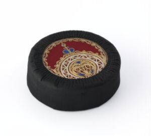  An ornate, round black sitting cushion with a richly decorated central design featuring gold and blue motifs on a red background, evoking a sense of luxury and detailed craftsmanship. Artistname and title are unknown.