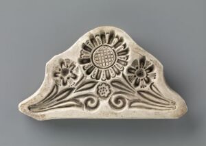  A triangular ceramic object with a curved top edge, adorned with a bas-relief design of a central sun-like motif and symmetric floral patterns on an off-white or light gray background. Artist name and title are unknown.