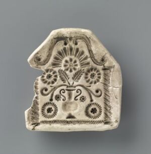  A small, pale cream-colored ceramic or stone fragment with a broken top right corner, showcasing a deeply embossed symmetrical design with central floral motif, mirrored stylized elements on sides, and various spiral and circular patterns, against a muted grey background.