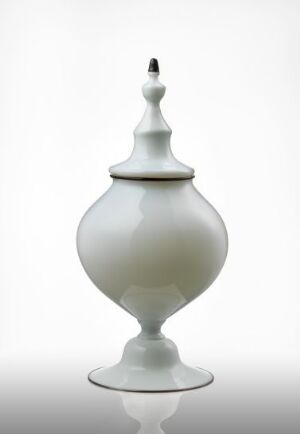  Translucent white glass vessel with a bulbous body and tapered neck, topped with a small, decorative finial, on a reflective surface against a white background. Artist's name and title unknown.