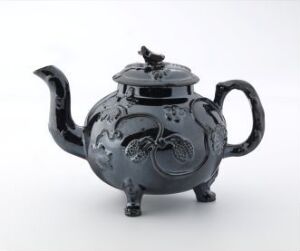  An intricately designed black teapot with floral patterns engraved, featuring a rounded body, a curved spout, an arched handle, and a decorative knob on the lid, set against a light gray background.