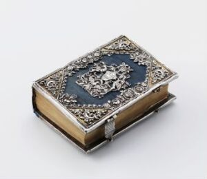  An ornate closed book with a deep blue cover featuring silver metallic designs, possibly velvet, with a central emblem and corner reinforcements, set against a plain light grey background.