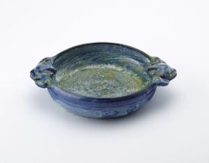  A shallow ceramic bowl with stylized animal head handles on the sides, featuring a rich blue exterior and a swirled green, yellow, and blue glaze inside, set against a light background.