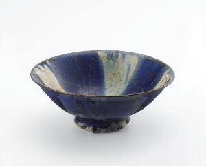  A small, footed ceramic bowl with a deep blue glaze and lighter blue-green streaks radiating from the center. The bowl has a handcrafted appearance and sits against a plain white background. Artistname and title are unknown.